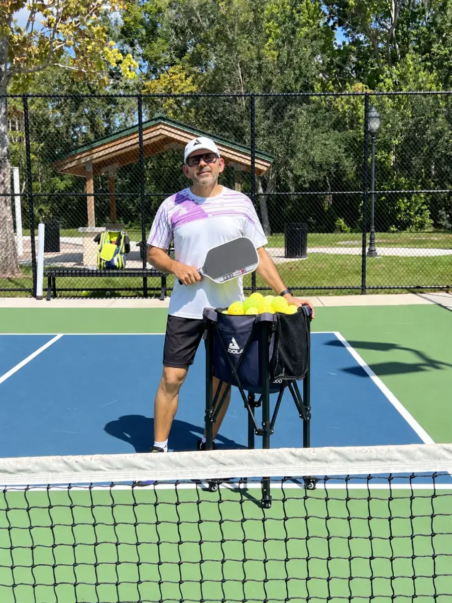 Coach Igor holding a pickleball paddle standing next to a basket of pickleballs on an outdoor court at Meadows Dog Park in New Port Richey, FL