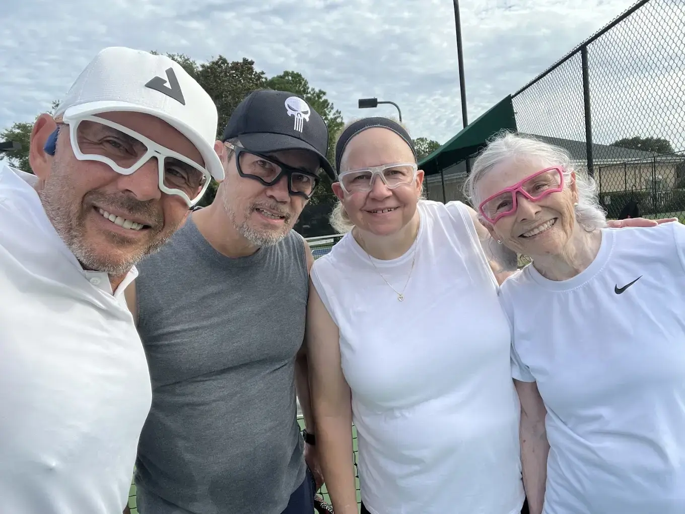 Four adults smiling at the camera on an outdoor pickleball court wearing sports eyewear, promoting pickleball lessons in New